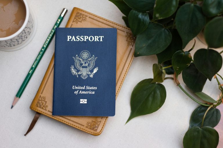 A U.S. passport rests on a small Bible surrounded by a cup of coffee, a sharpened pencil and trailing pothos leaves.