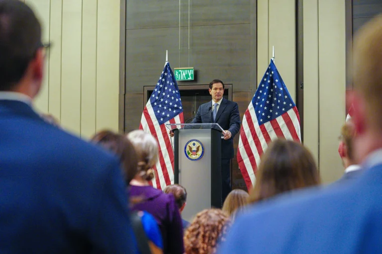 Marco Rubio standing at a podium, speaking with U.S. flags on both sides and a few people seated in the audience.