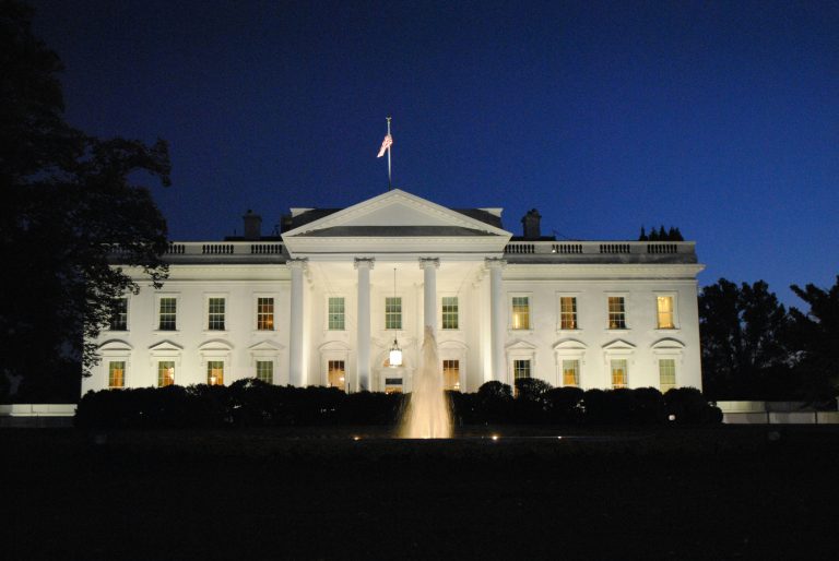 Front view of the White House at night, fully illuminated, with a dark sky in the background.
