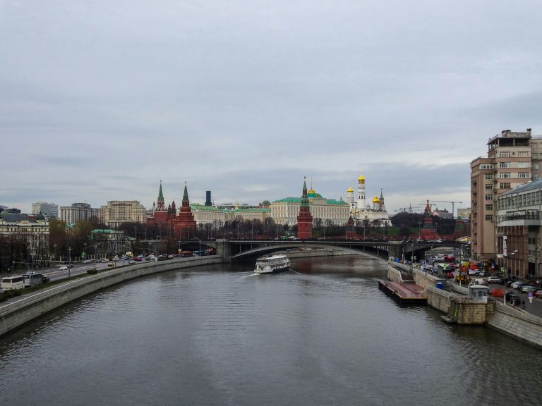 A view of the Moscow Kremlin along the Moskva River under a cloudy sky, with its historic red brick walls, golden-domed churches, and towers reflected in the water.
