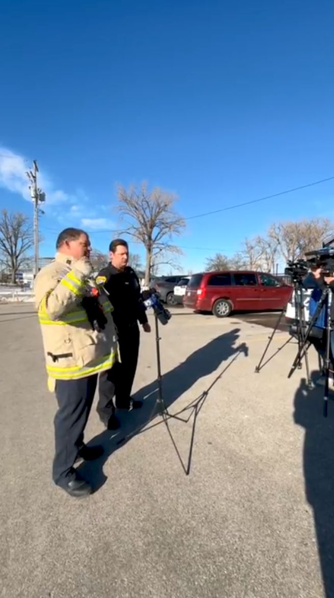 A firefighter and a police officer speak to media microphones and cameras during an outdoor press conference, with vehicles and trees under a clear blue sky.