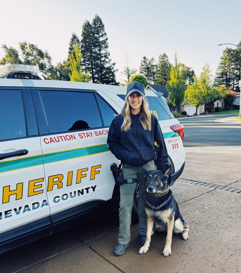Deputy Kayla Ramos standing next to a Nevada County Sheriff patrol vehicle with K9 Riggs sitting by her side.