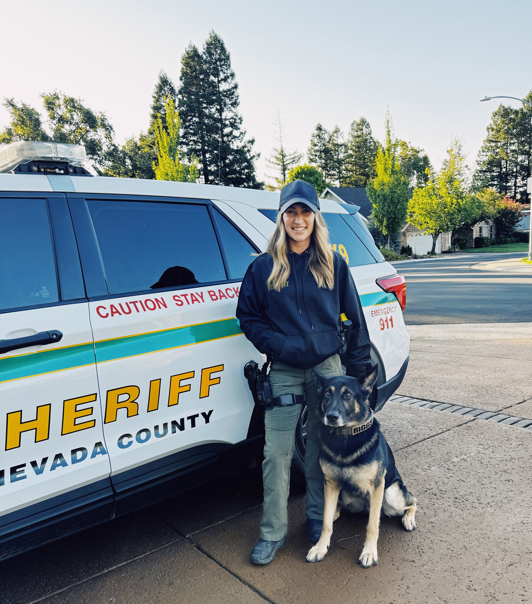 Deputy Kayla Ramos standing next to a Nevada County Sheriff patrol vehicle with K9 Riggs sitting by her side.