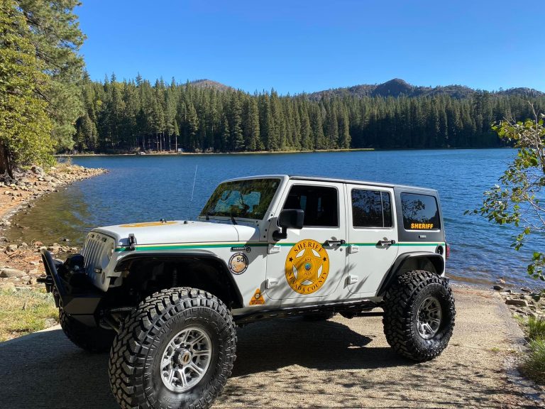 Nevada County Sheriff marked 4-door white Jeep with large off-road tires and custom bumper parked on a paved shore next to a calm lake, with dense pine forest and mountains under a blue sky.