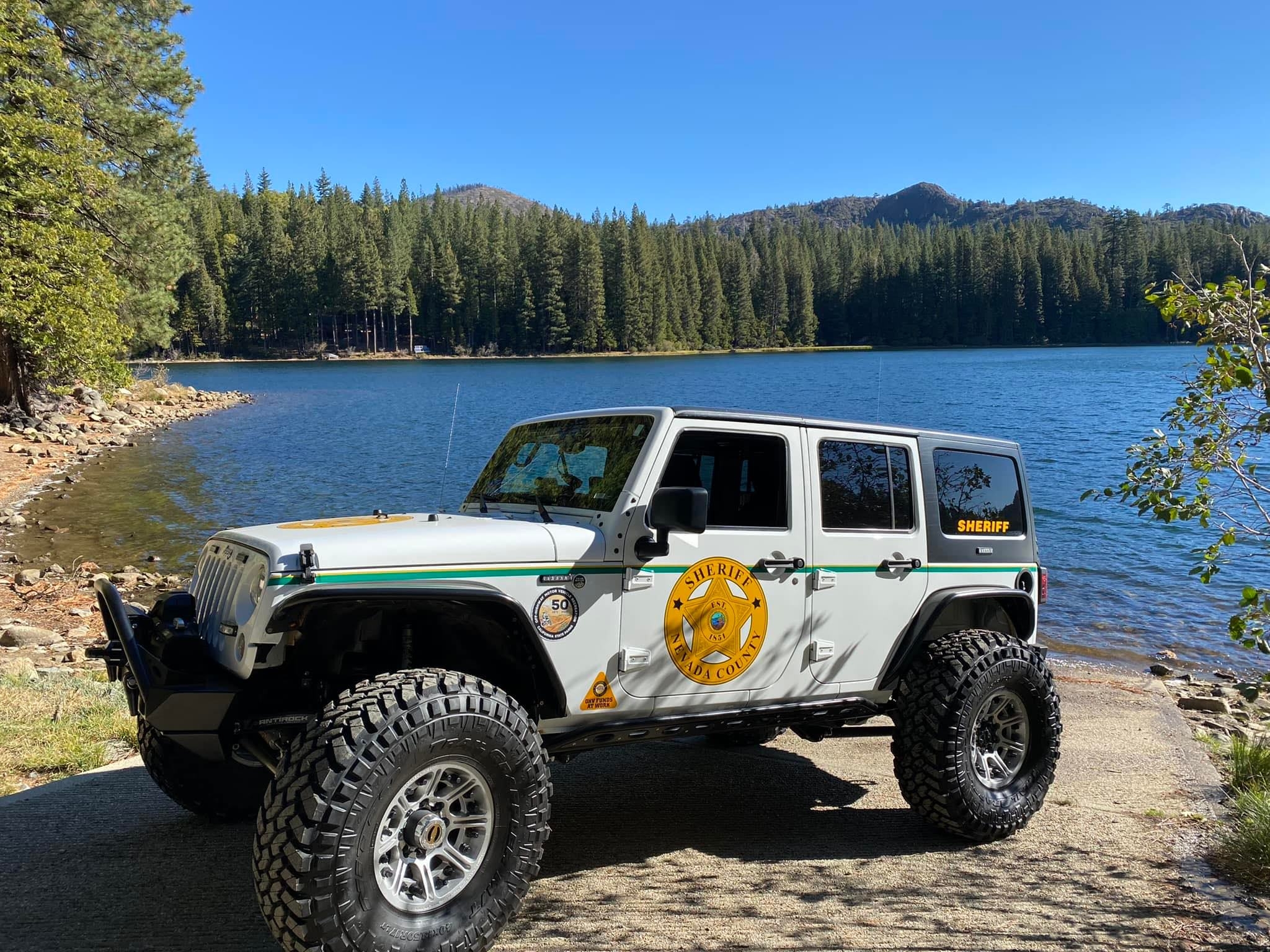 Nevada County Sheriff marked 4-door white Jeep with large off-road tires and custom bumper parked on a paved shore next to a calm lake, with dense pine forest and mountains under a blue sky.