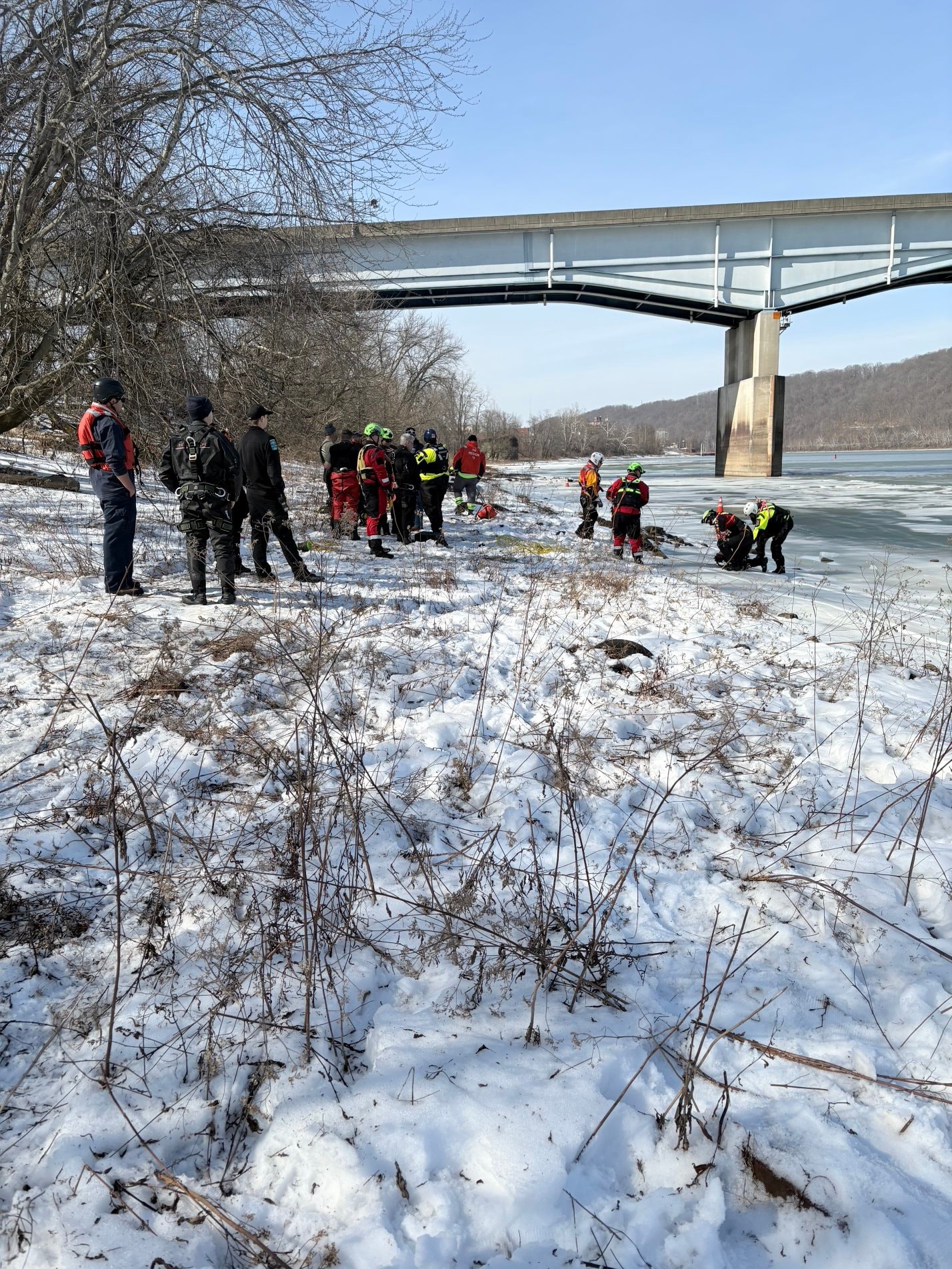 Search and rescue teams in high-visibility gear standing on a snowy riverbank near an iced-over Monongahela River under a bridge.