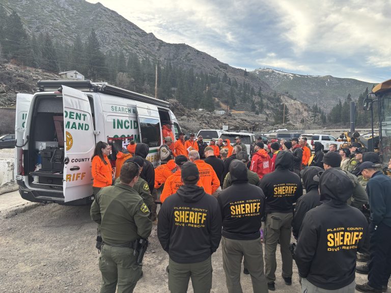 A large group of search and rescue personnel from the Placer County and Nevada County Sheriff's offices gathered for a briefing next to a white mobile incident command vehicle in a mountainous region.
