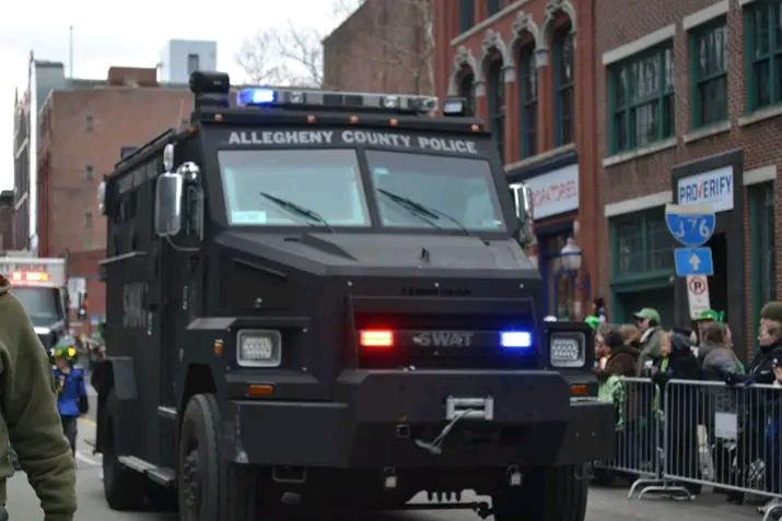 An Allegheny County Police SWAT armored vehicle driving through a city street lined with crowds behind metal barricades.