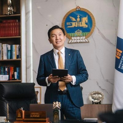 A man in a blue pinstriped suit, gold tie, and white shirt stands in an office holding a tablet. A Mongolian Soyombo emblem and a flag are visible.