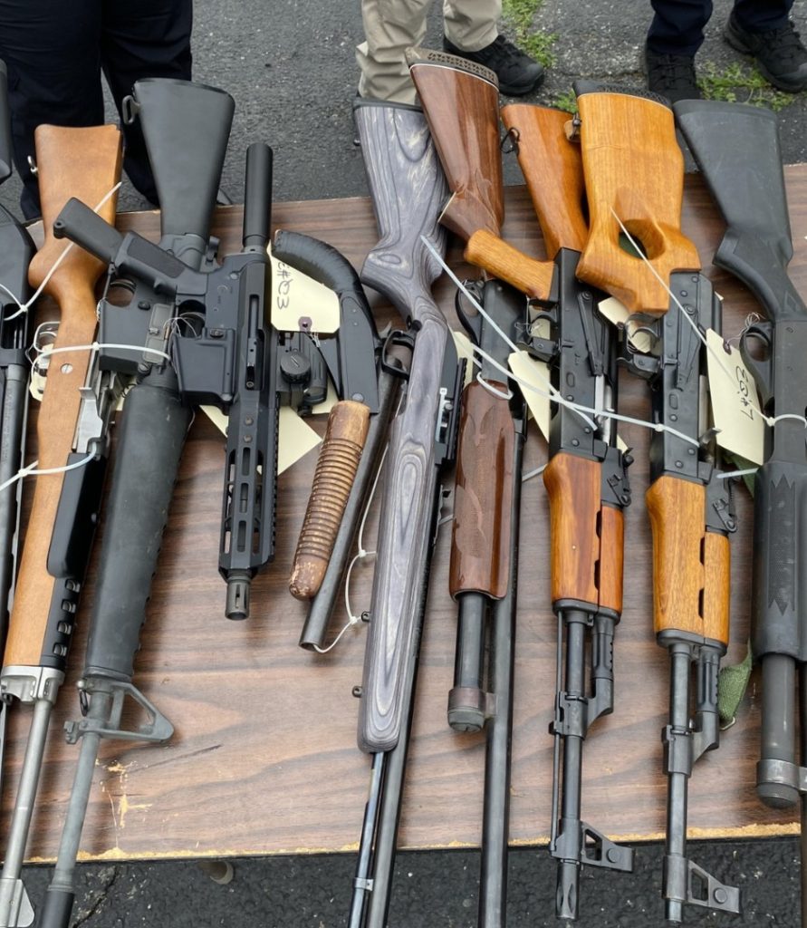 A top-down view of several long guns, including wooden-stock rifles and tactical-style assault weapons, tagged with yellow labels and secured with white zip ties on a wooden table.