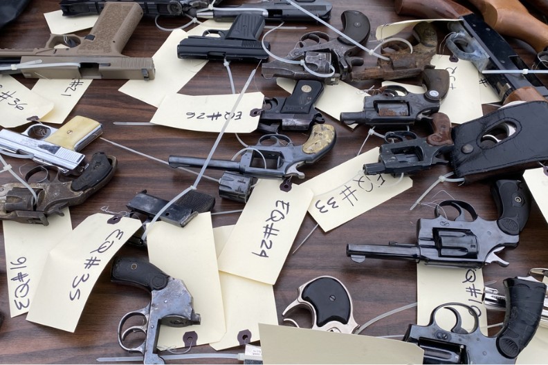 A close-up view of several handguns and revolvers laid out on a table, secured with zip ties and labeled with yellow inventory tags.
