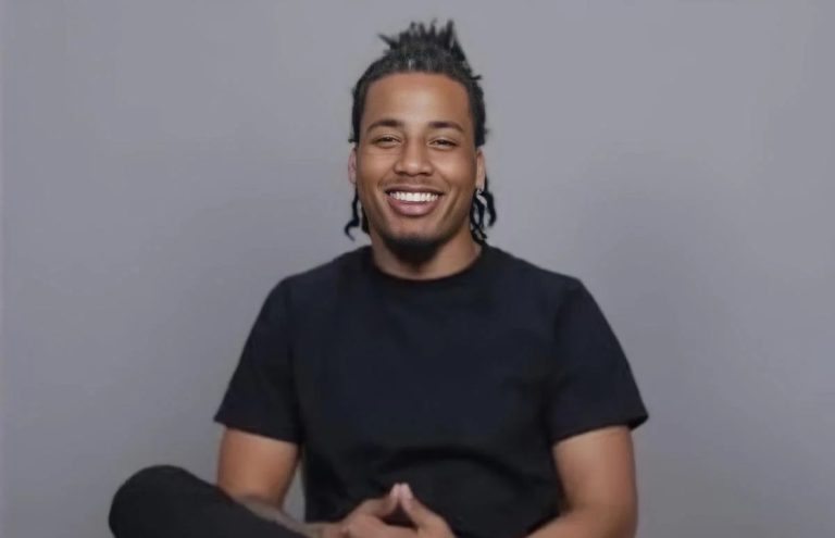 Portrait of 28-year-old Hadari Darden, a Black man with dreadlocks, smiling in front of a neutral grey studio background.