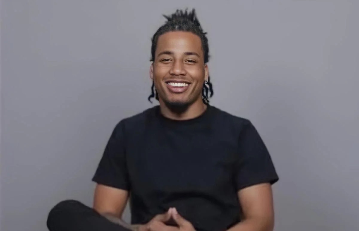 Portrait of 28-year-old Hadari Darden, a Black man with dreadlocks, smiling in front of a neutral grey studio background.