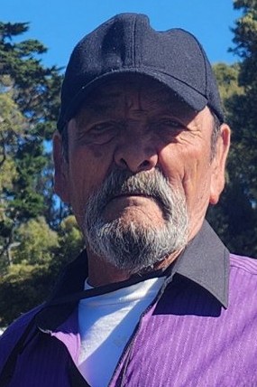 A headshot of a 76-year-old Latino male with a gray mustache and beard, wearing a black hat, a white t-shirt, and a purple collared shirt. He is outdoors with green trees and a blue sky in the background.