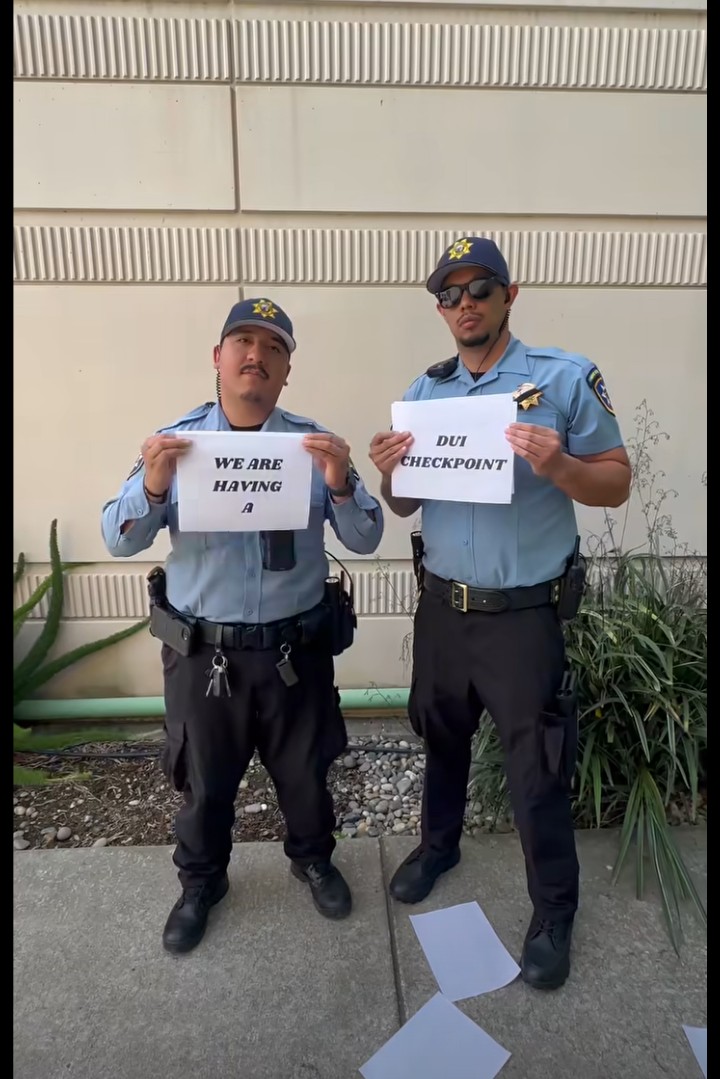 Two Redwood City police officers in light blue uniforms hold signs reading "WE ARE HAVING A" and "DUI CHECKPOINT" while standing outdoors.