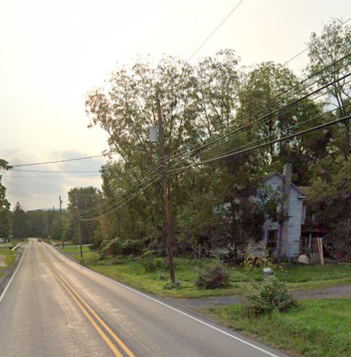 A two-story residential house situated along a two-lane paved road with utility lines and dense trees in the background under a pale sky.