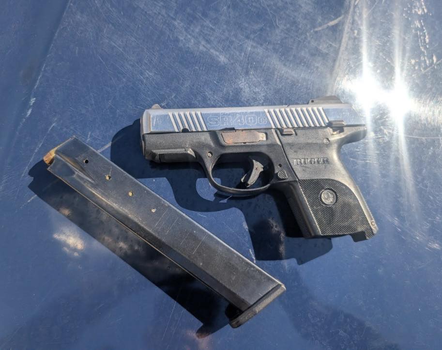 A Ruger SR40c semi-automatic handgun with a silver slide and black frame rests on a dark surface next to a detached extended capacity magazine." https://gemA Ruger SR40c semi-automatic handgun with a silver slide and black frame rests on a dark surface next to a detached extended capacity magazine.