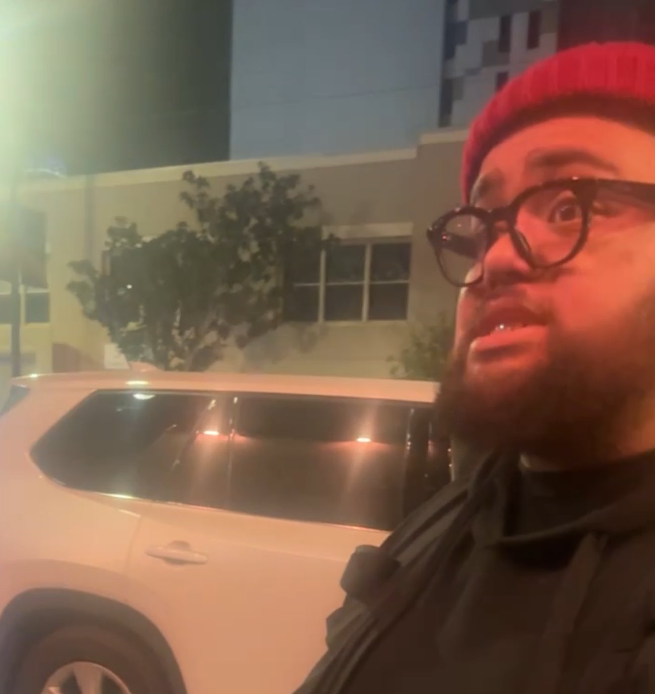 A male suspect with a beard, wearing black-rimmed glasses, a black hooded sweater, and a red beanie cap, standing on a city street at night.