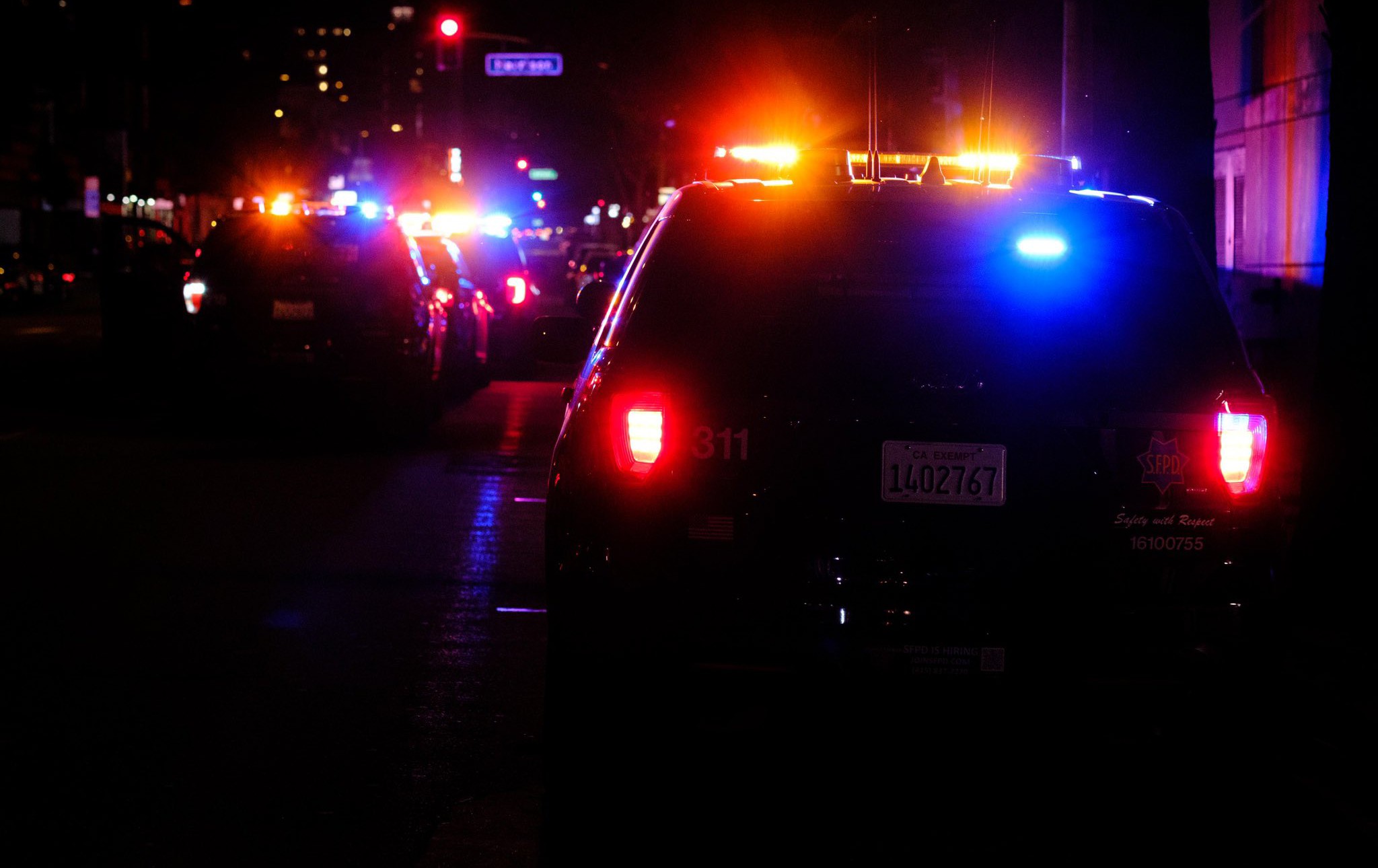 Multiple San Francisco Police Department patrol cars with emergency lights activated on a city street. The rear of a vehicle with license plate 1402767 is prominent.