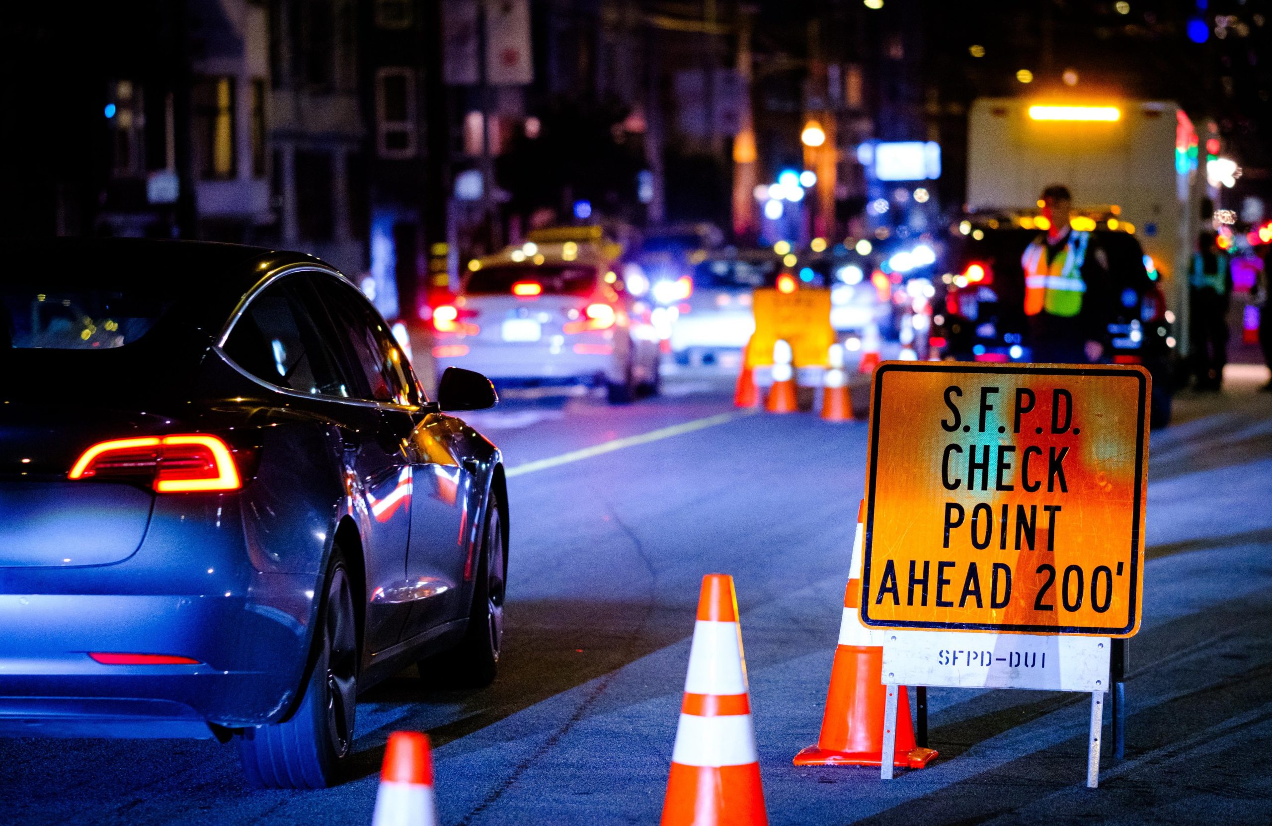 An orange traffic sign reading 'S.F.P.D. CHECKPOINT AHEAD 200 FT.' at night, with a dark car approaching and a police officer in a safety vest visible in the background.