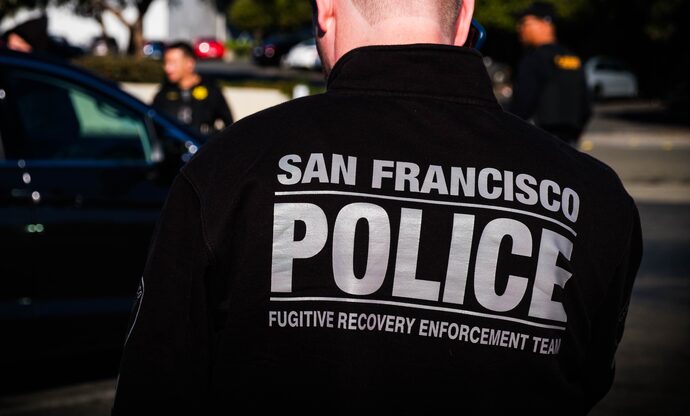 Rear view of an SFPD officer in a black jacket featuring the text "SAN FRANCISCO POLICE" and "FUGITIVE RECOVERY ENFORCEMENT TEAM" in white letters. The background shows blurred officers and vehicles.