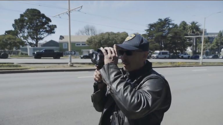 A close-up shot of a San Francisco Police Department Traffic Enforcement Officer in a leather jacket and sunglasses, holding and looking through a handheld speed laser device (LIDAR) near a multi-lane road.