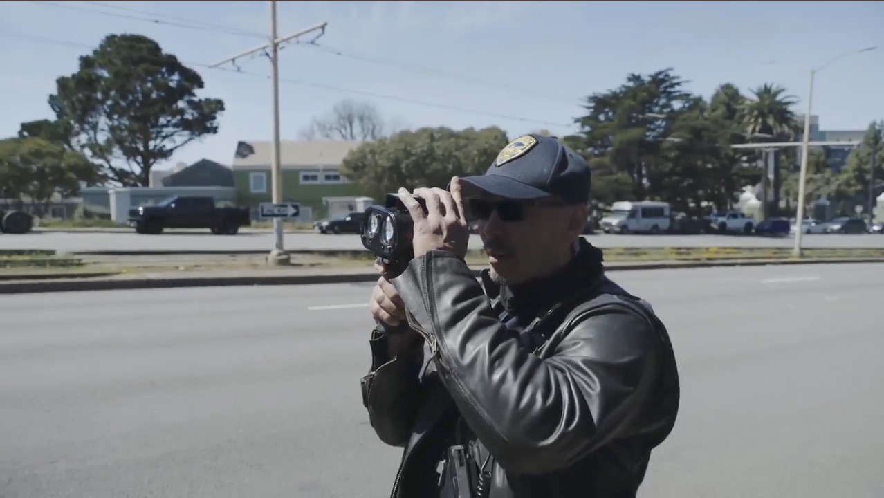 A close-up shot of a San Francisco Police Department Traffic Enforcement Officer in a leather jacket and sunglasses, holding and looking through a handheld speed laser device (LIDAR) near a multi-lane road.
