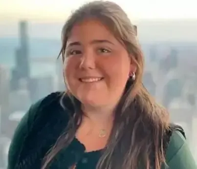 A portrait of Sheridan Gorman, a smiling young woman with long brown hair, wearing silver hoop earrings and a green top, with a blurred Chicago skyline visible in the background.