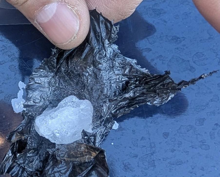 A close-up view shows a person’s thumb holding open a piece of black plastic material containing a crystalline, white substance suspected to be methamphetamine.