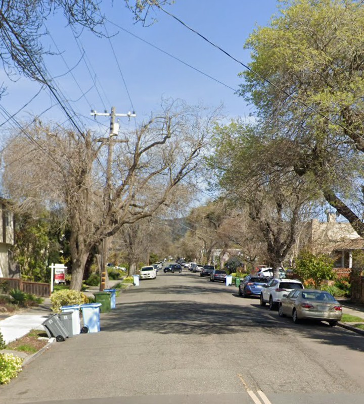 A residential street in Berkeley, California, showing parked cars, utility poles, and large trees lining the roadway under a clear sky.