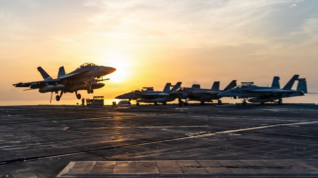 An F/A-18 Super Hornet lands on the flight deck of an aircraft carrier at sunset, with several other fighter jets parked nearby.