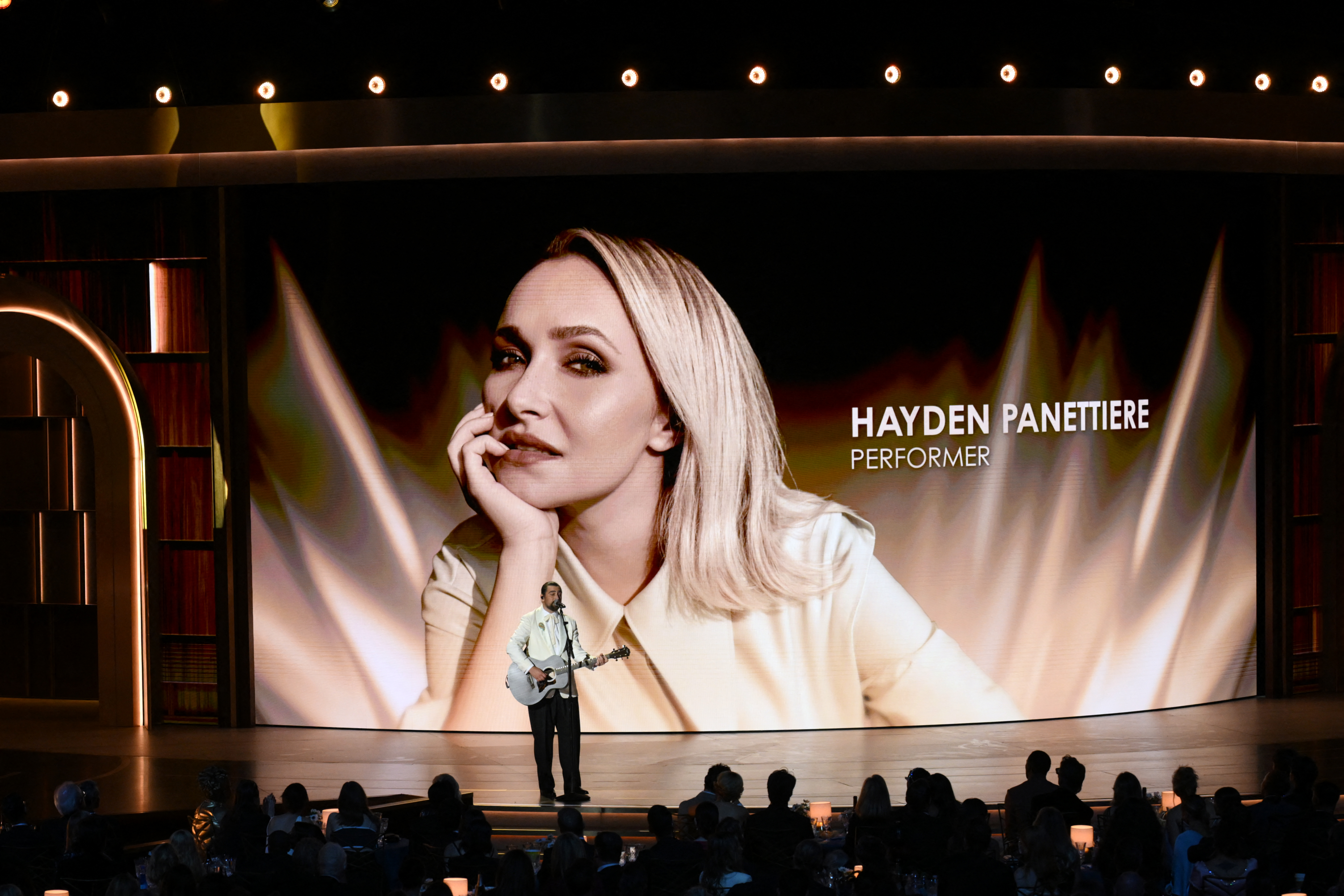 A wide shot of a stage featuring a large digital screen displaying a portrait of late US actress Hayden Panettiere with the text "HAYDEN PANETTIERE PERFORMER." Below the screen, a lone male musician stands holding an acoustic guitar, facing an audience seated in a dark, dimly lit venue under a row of bright overhead lights.