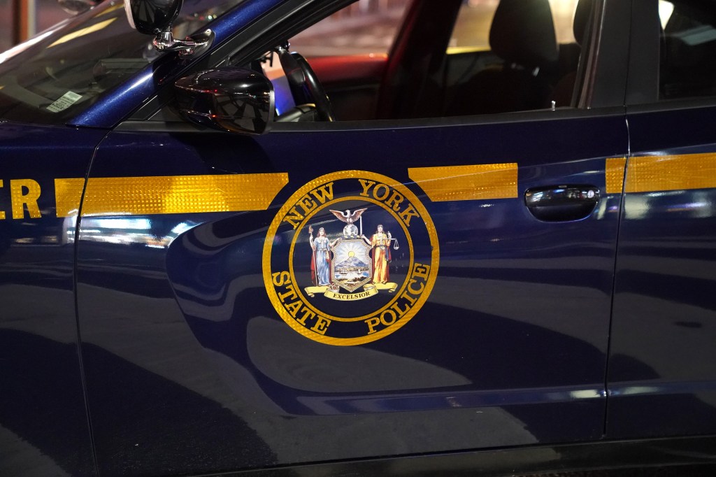 A close-up shot of the side door of a dark blue New York State Police cruiser. The vehicle features a prominent circular gold and blue emblem with the words "New York State Police" surrounding a detailed state seal. A reflective yellow stripe runs horizontally across the body panel near the top, and the side mirror is visible in the upper left.