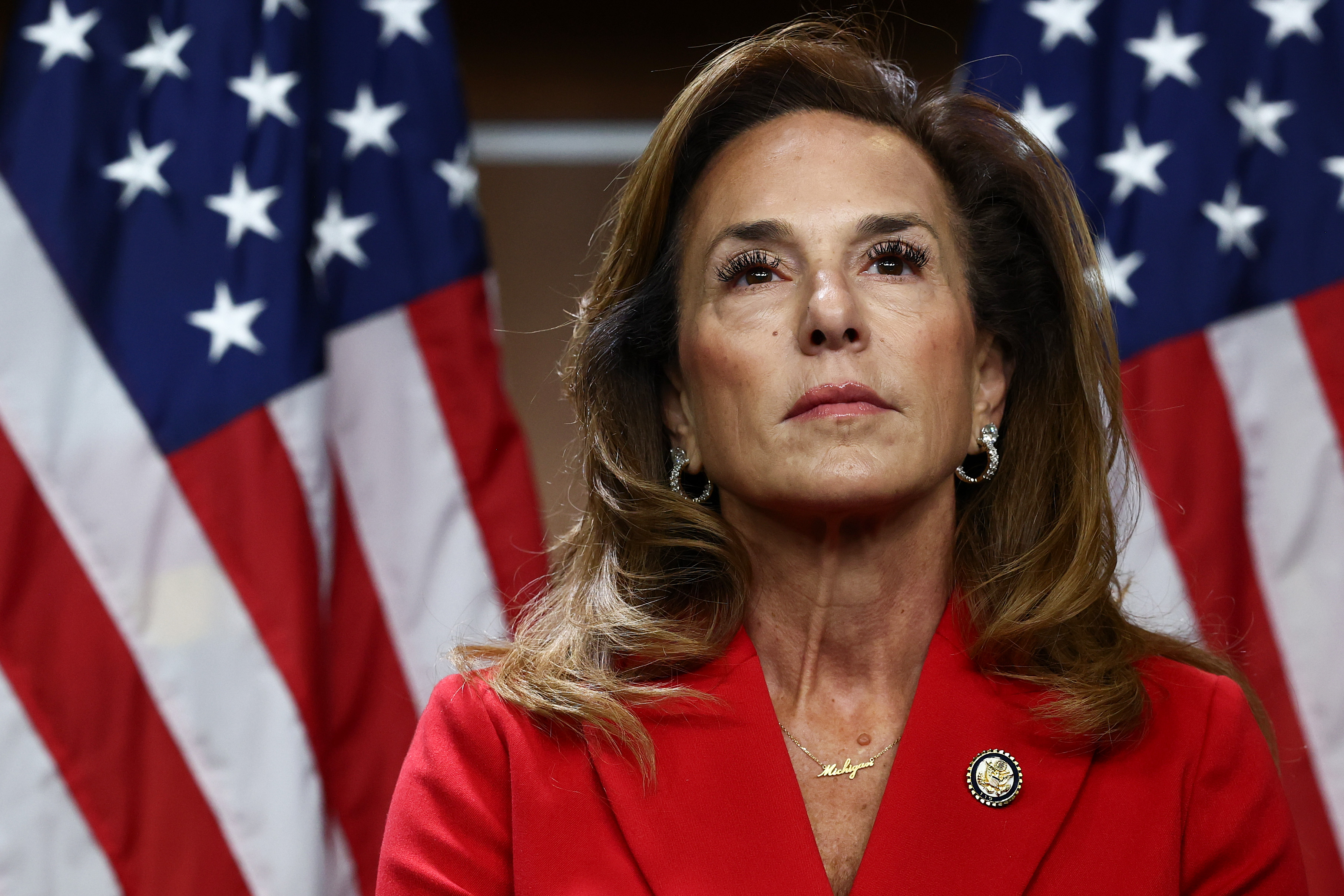 A medium close-up shot of U.S. Representative Lisa McClain standing before two American flags. She wears a vibrant red blazer pinned with a small brooch and a delicate gold necklace, complemented by dangling earrings. With shoulder-length brown hair and a composed expression, she gazes slightly upward and away from the camera.